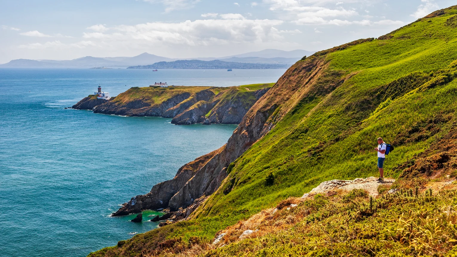 A person standing on a trail on a hill overlooking the coast