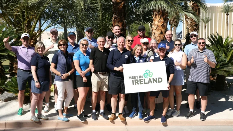 Group of attendees posing outdoors at an IMEX 2025 event, holding a ‘Meet in Ireland’ sign, with palm trees and a venue building in the background.