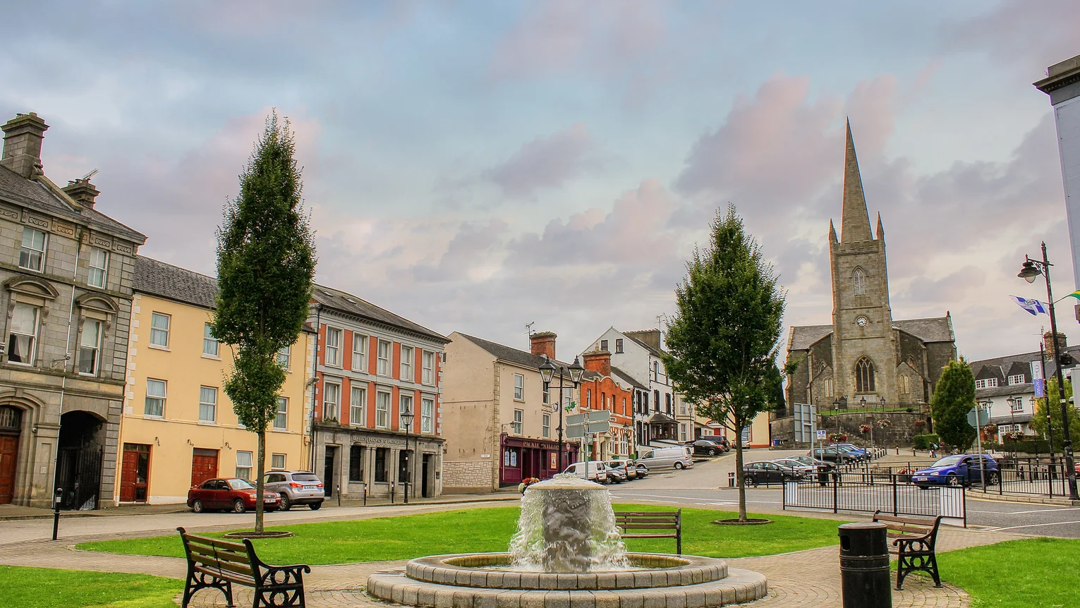 A fountain in a town centre with trees and buildings in the background