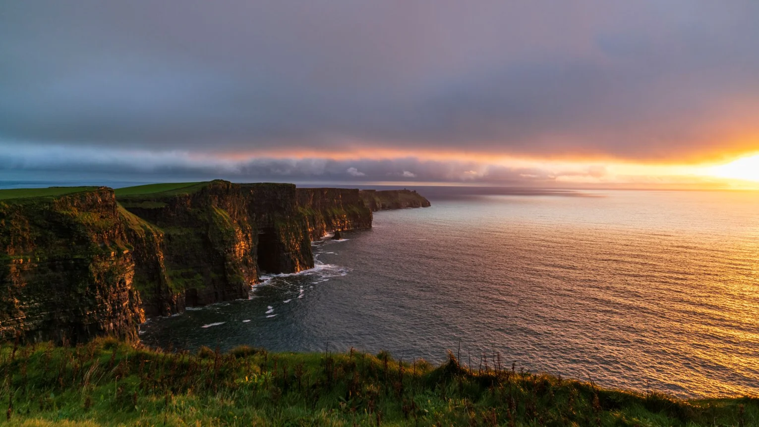 Sunset light over the Cliffs of Moher in Co. Clare, with rugged sea cliffs overlooking the Atlantic Ocean beneath dramatic clouds.