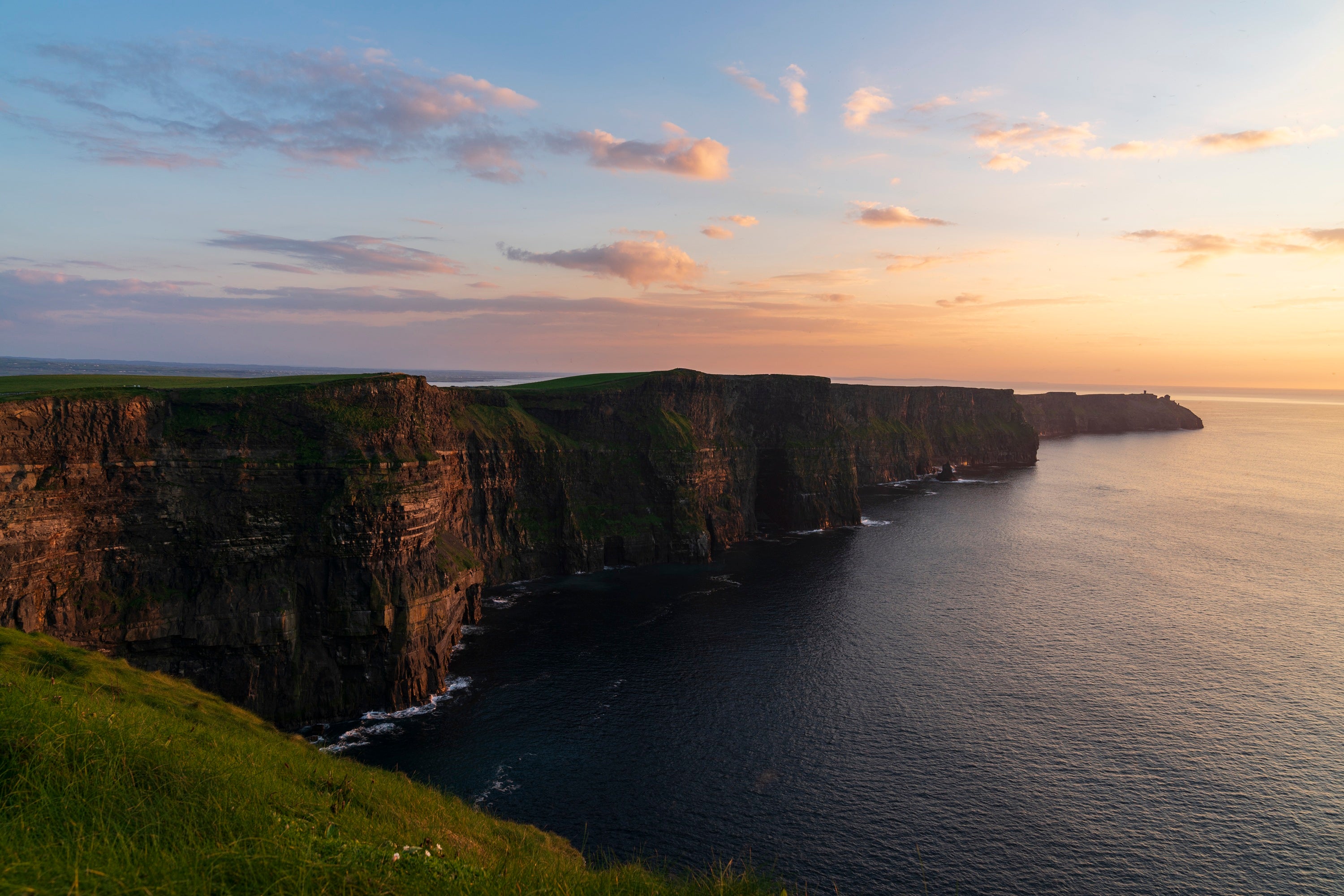A cliff above water with grass and a sunset with Cliffs of Moher in the background
