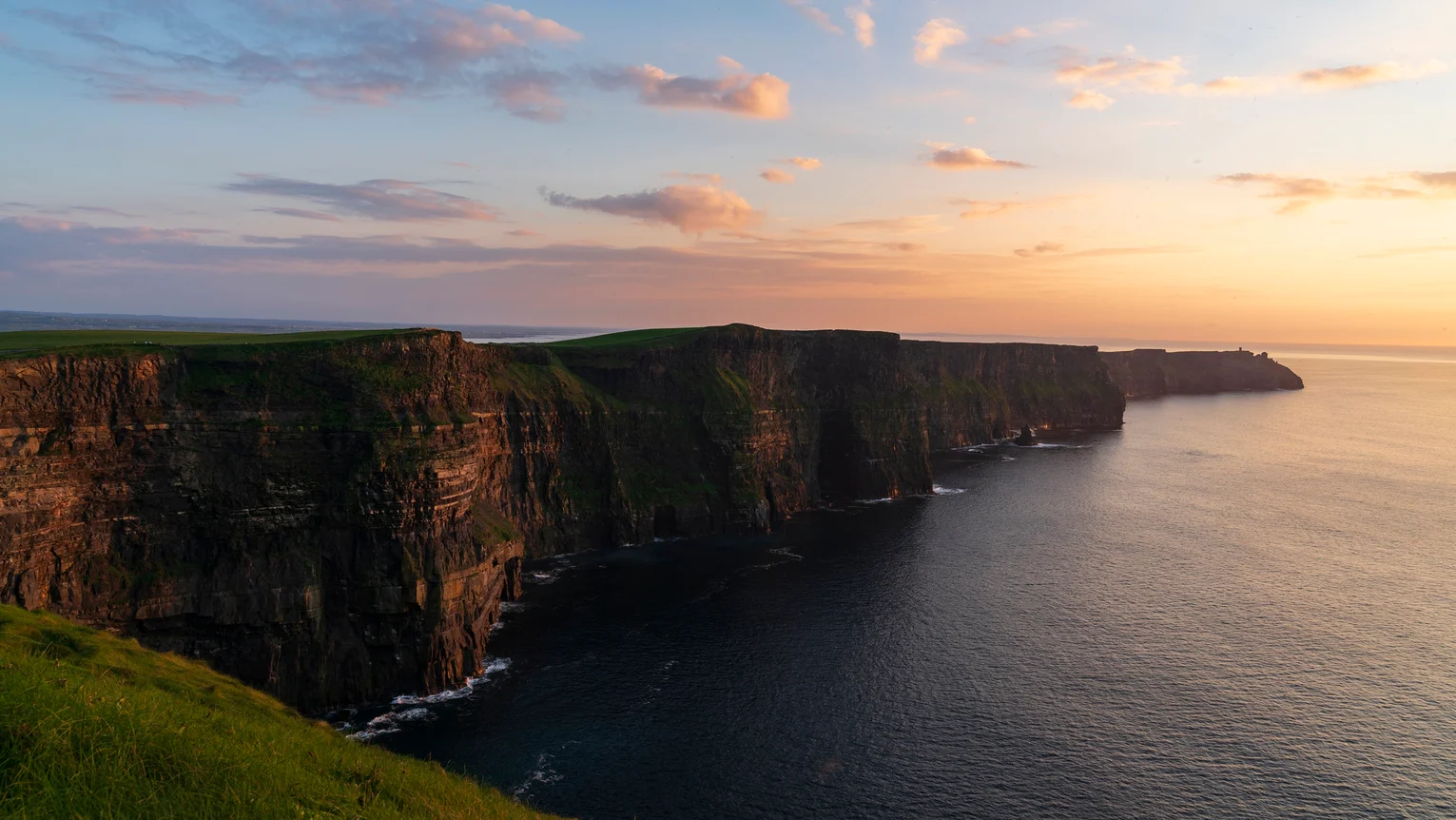 A cliff above water with grass and a sunset with Cliffs of Moher in the background