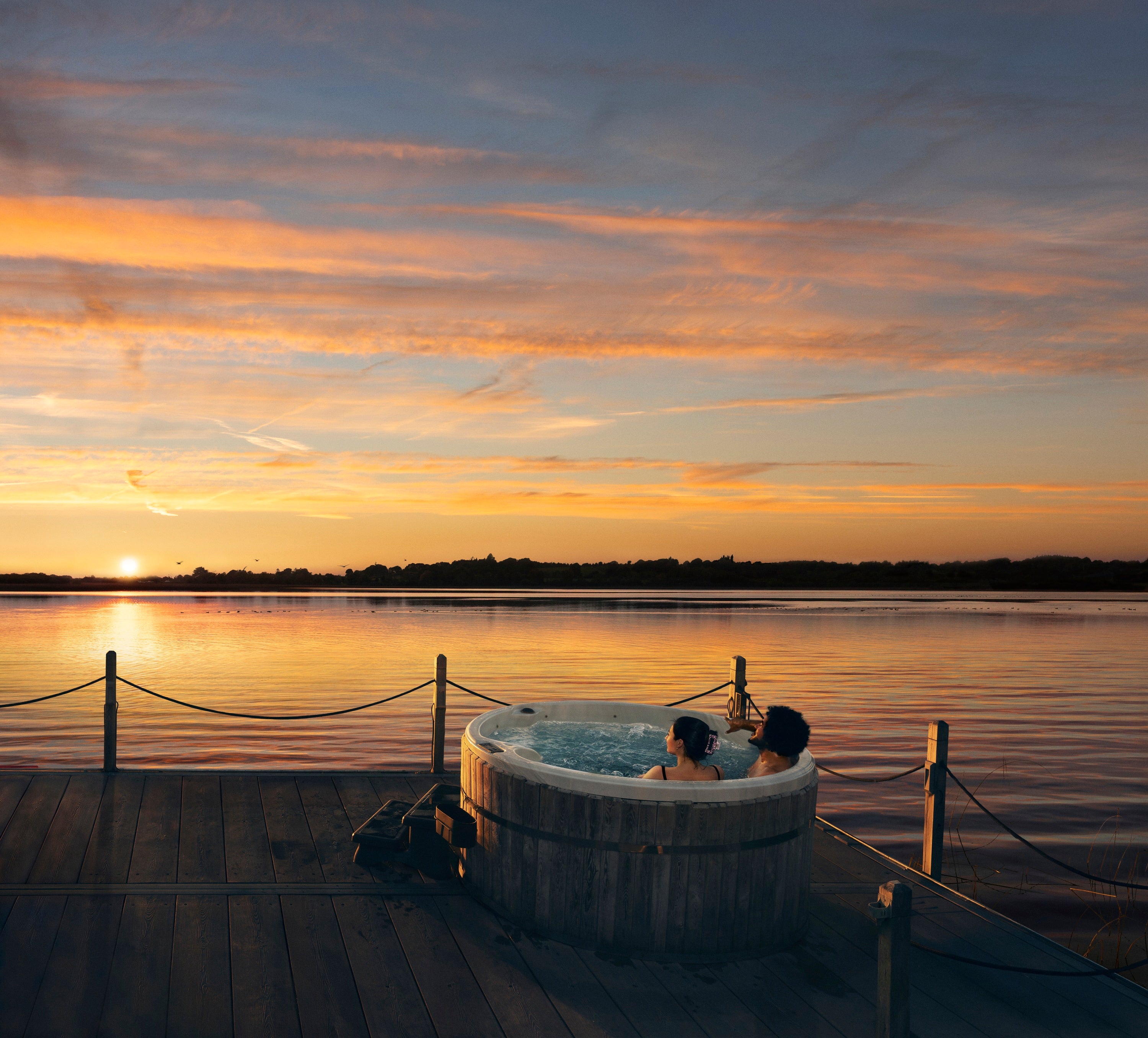 A couple of people in a hot tub on a dock with a sunset